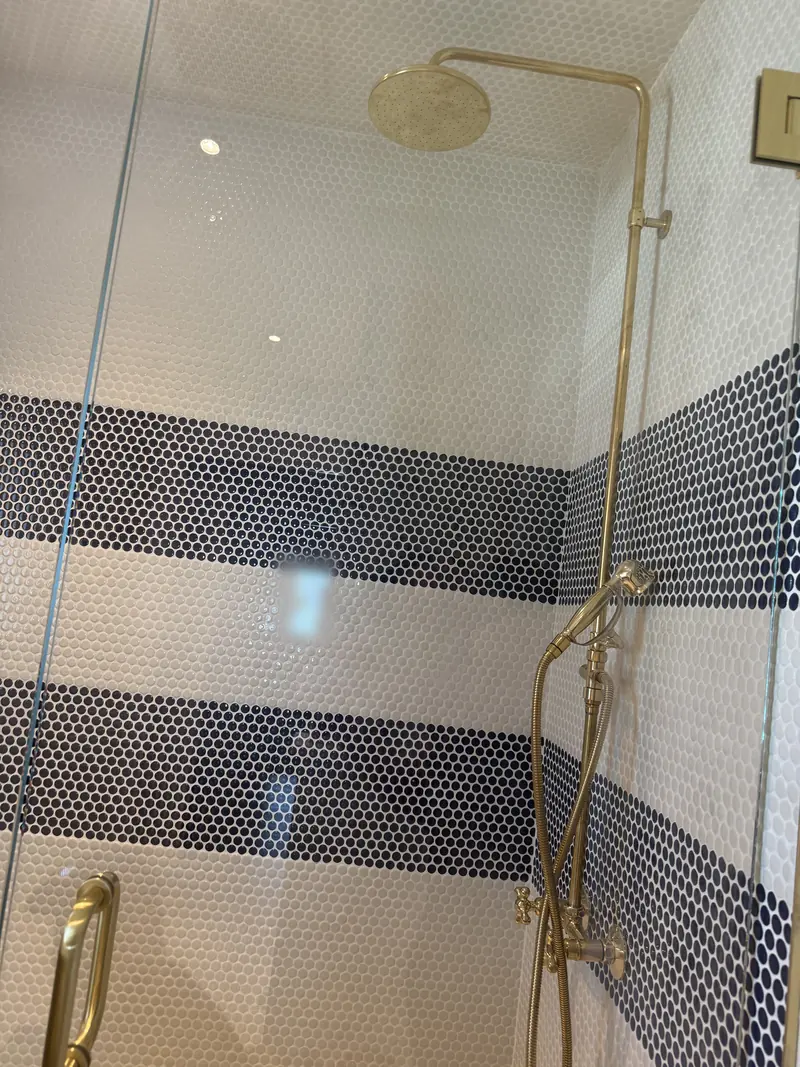 Shower with a brass rain head, exposed brass riser and handheld shower set in navy and white striped penny tile, behind a glass door