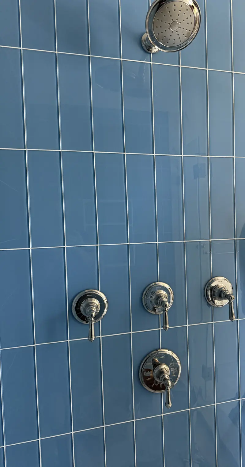 Blue glass tile shower wall with a showerhead, three polished nickel valves and a round valve trim below