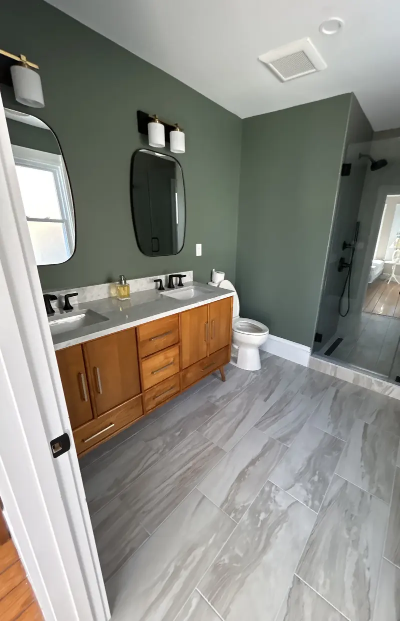 Bathroom with sage green walls, a wood double vanity with matte black faucets, a toilet and a frameless glass shower
