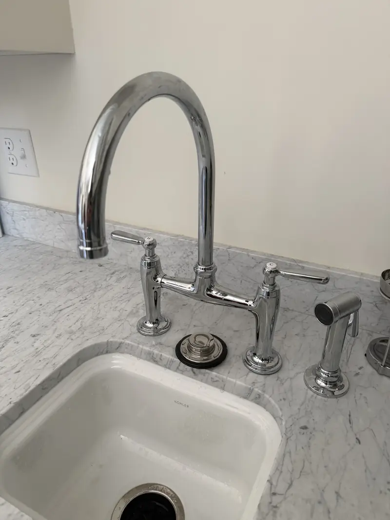 Polished chrome bridge faucet with lever handles and a side sprayer over a white undermount sink on a marble counter